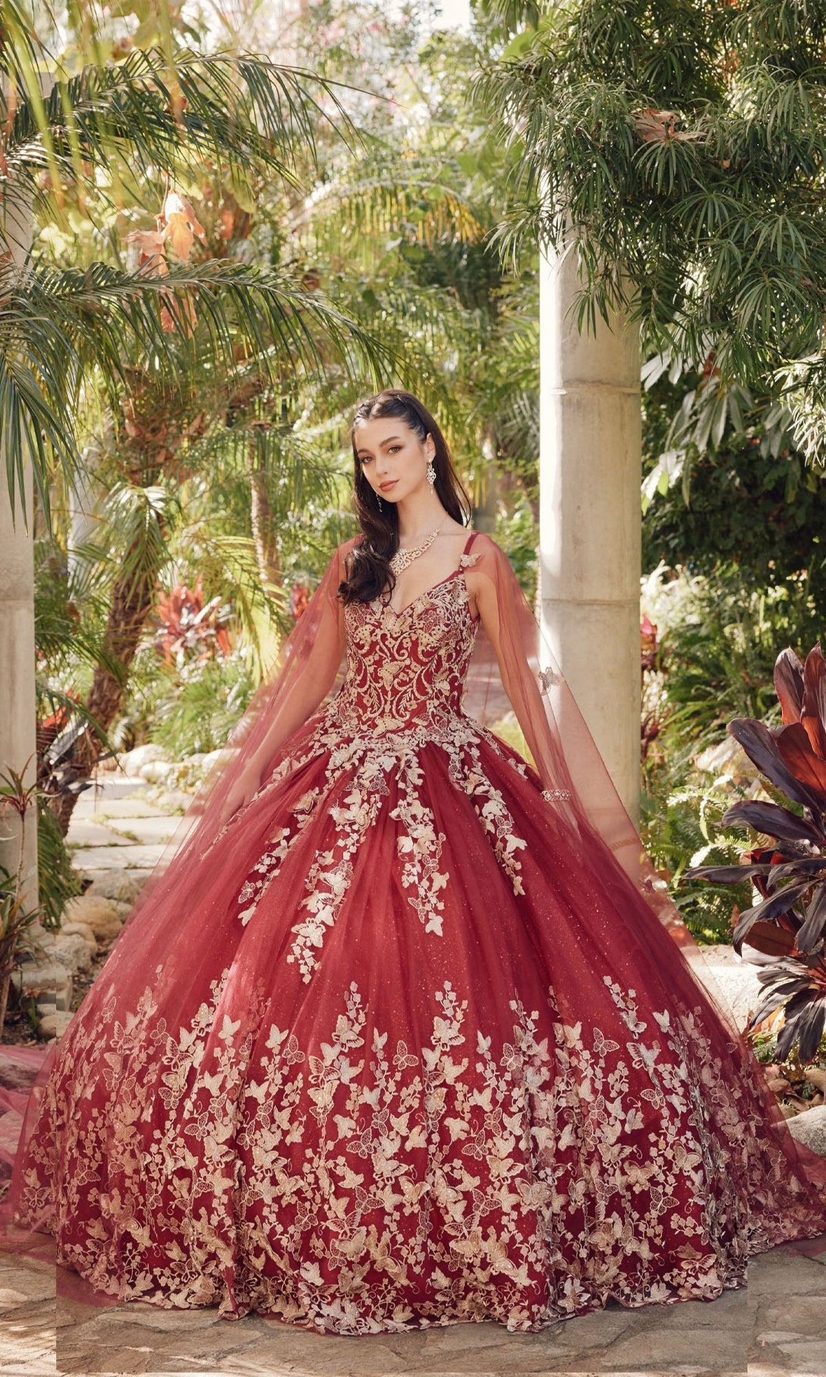 In a lush garden, a woman wears the Juliet 1442 Quinceañera Dress with gold floral embroidery, tulle accents, and a sheer cape, surrounded by greenery and stone columns.